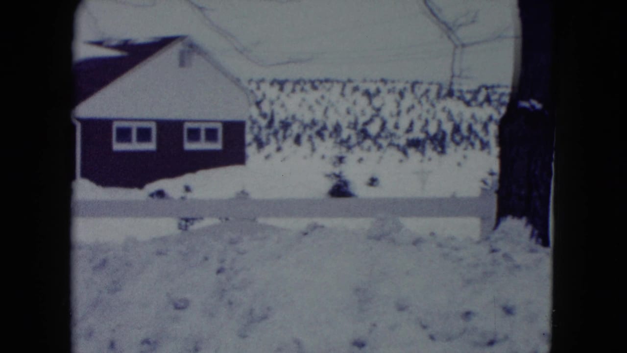 Snowy yard and neighboring house seen behind a low white fence.