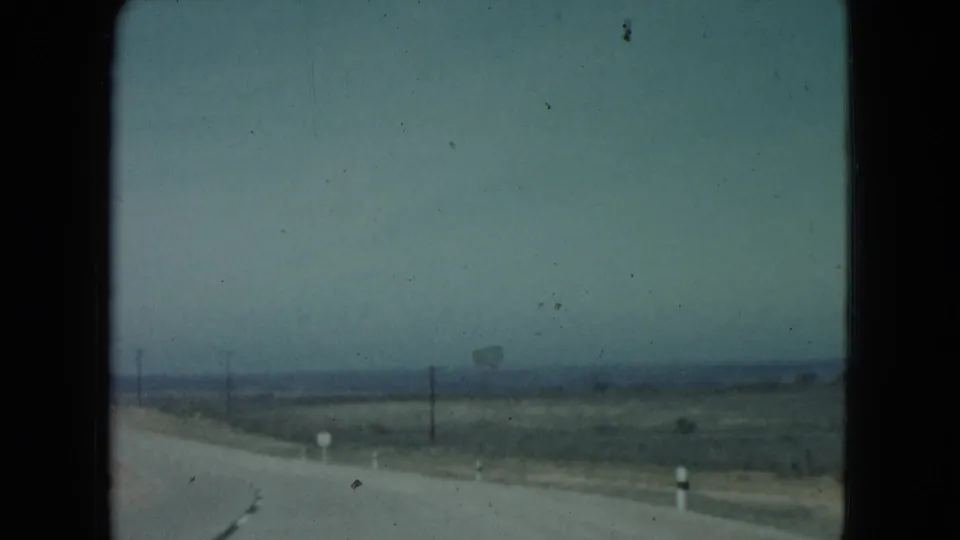 Nevada desert road seen from a moving vehicle, with roadside posts, utility poles, and low hills in the distance.