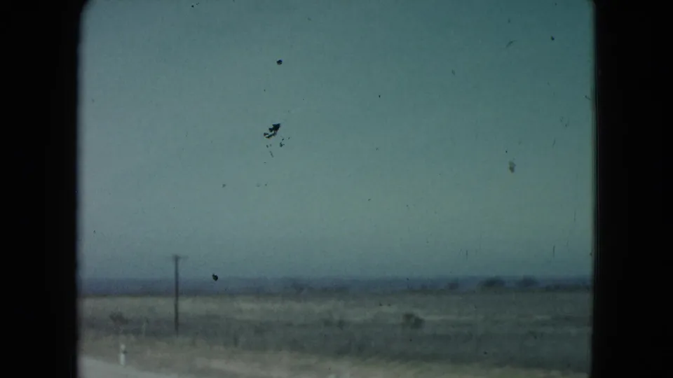 Soft 8mm view of Nevada desert flats, a roadside pole, and a low horizon as the car moves past the landscape.