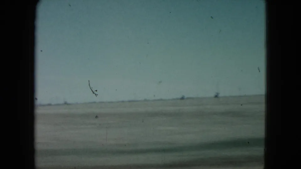 1960 Nevada 8mm frame showing open desert, a low horizon, pale sky, and faint distant structures seen from a moving viewpoint.