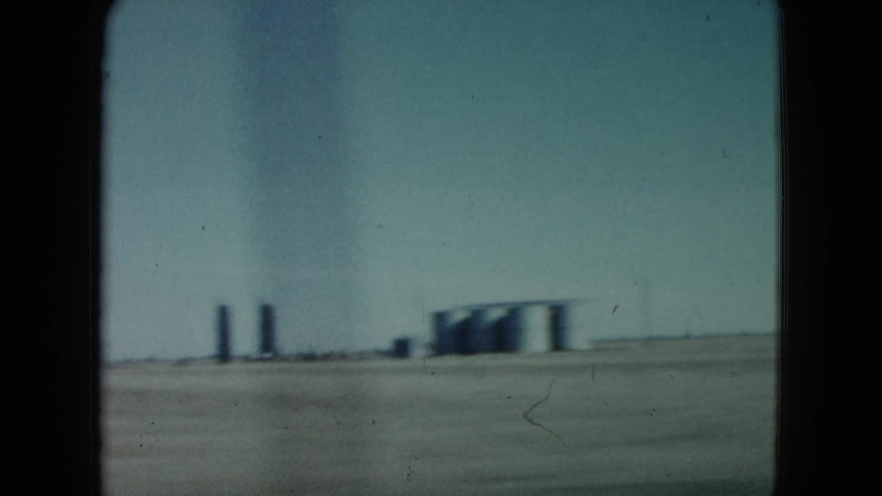 Distant industrial tanks and vertical structures across a flat desert foreground.