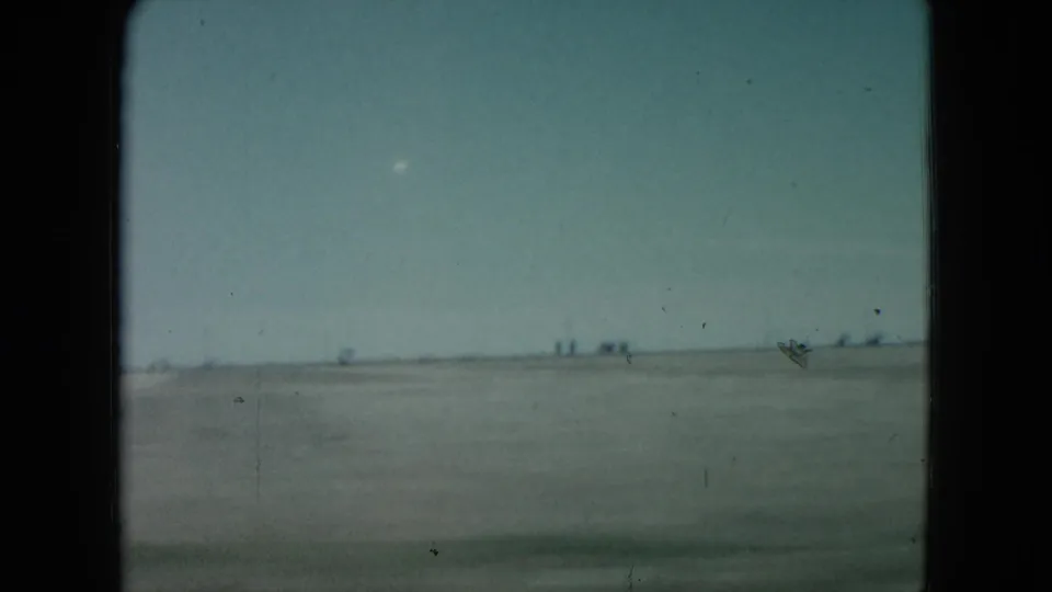 1960 Nevada 8mm frame of a wide desert plain, pale sky, distant horizon marks, dust, scratches, and motion blur.