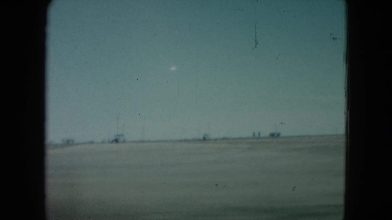 1960 Nevada 8mm frame showing a flat desert horizon, faint distant structures, and a small bright white object in the pale sky.