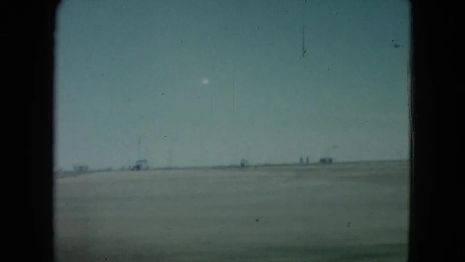 1960 Nevada 8mm frame showing a flat desert horizon, faint distant structures, and a small bright white object in the pale sky.