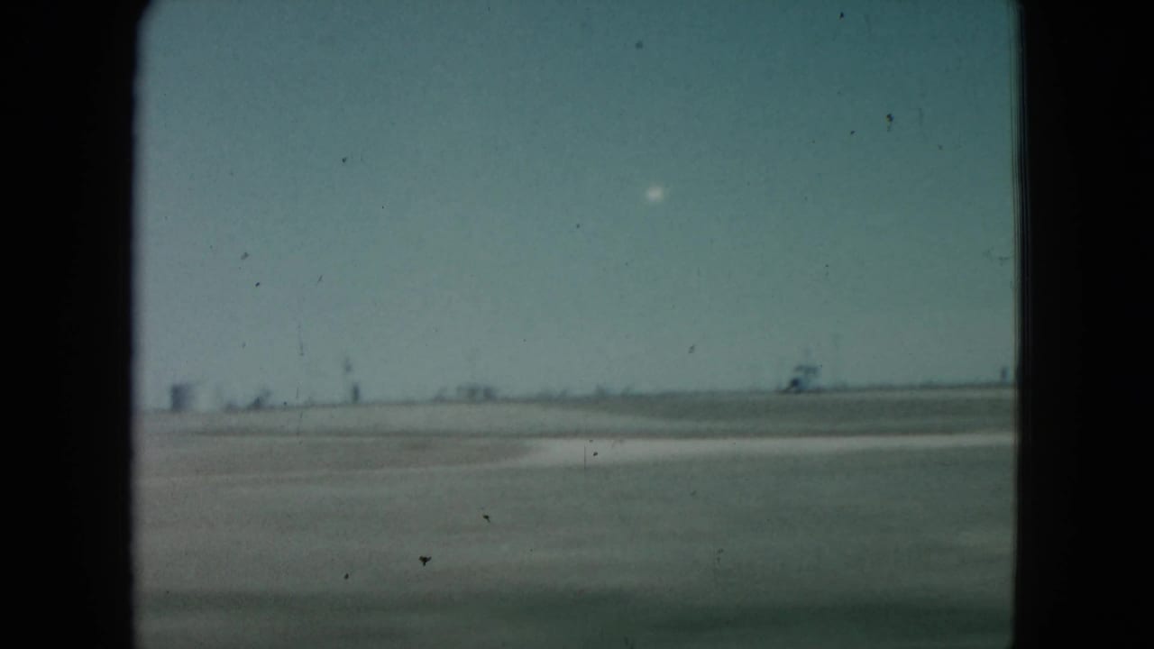 1960 Nevada 8mm frame with open desert terrain, a bright white object above the horizon, and distant low structures softened by film grain.