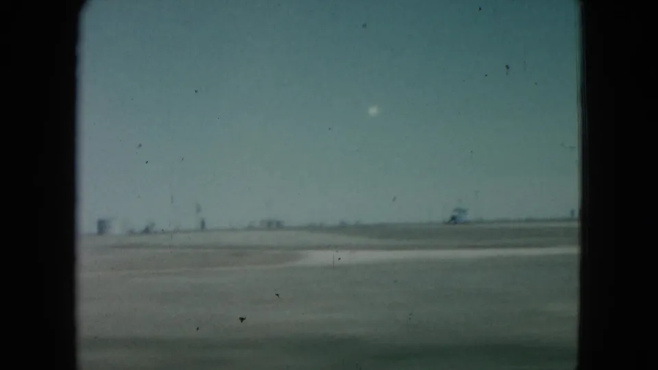 1960 Nevada 8mm frame with open desert terrain, a bright white object above the horizon, and distant low structures softened by film grain.