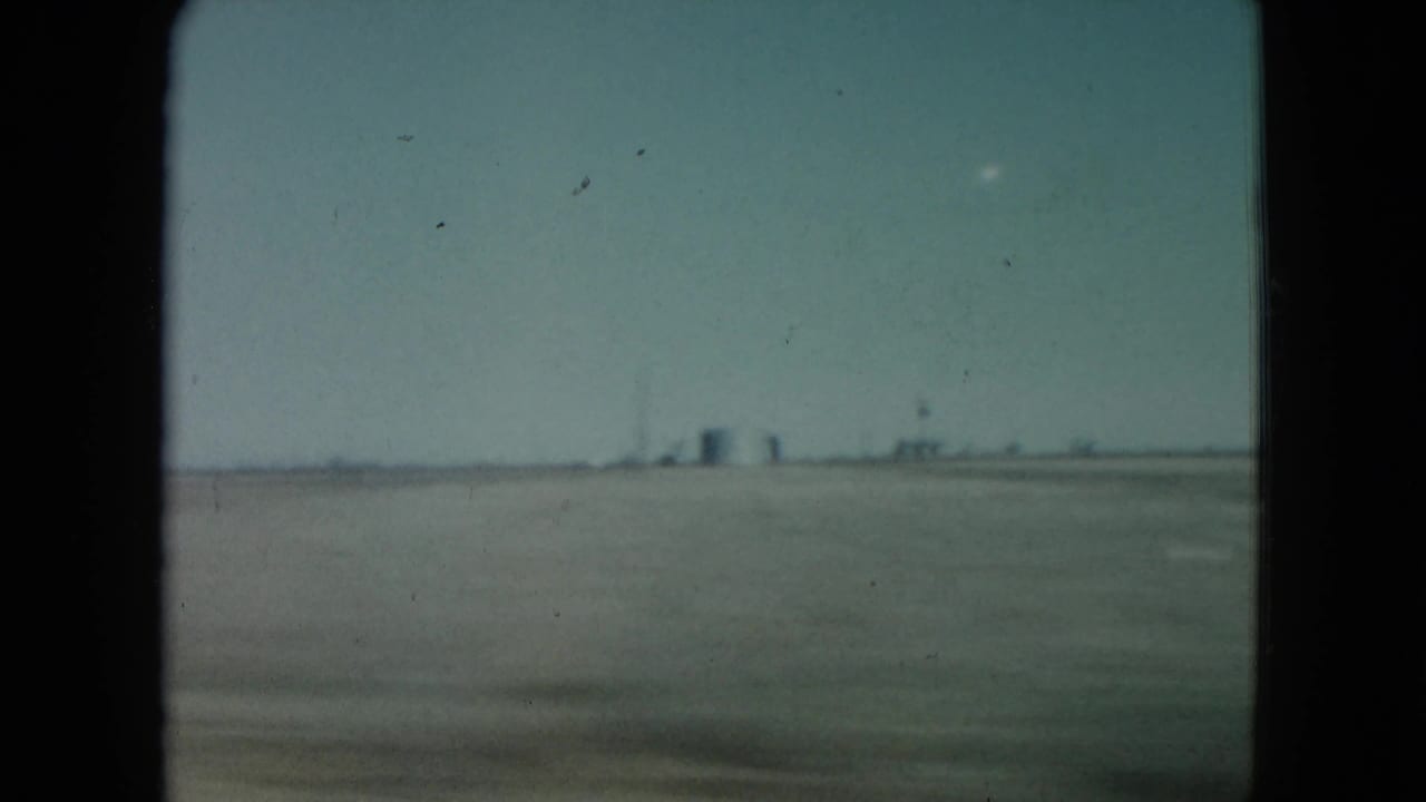 1960 Nevada 8mm frame showing a broad desert foreground, faint industrial structures near the horizon, and a small pale object in the sky.