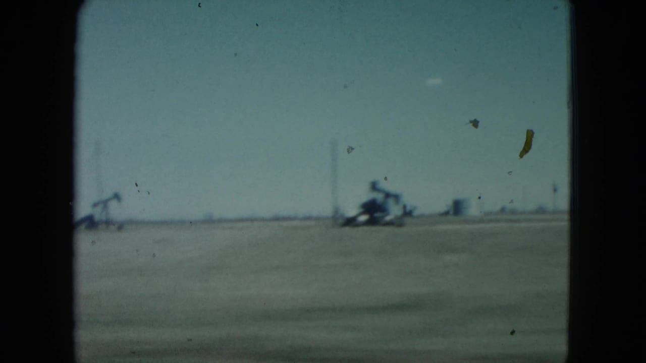 1960 Nevada 8mm frame showing flat desert terrain, visible pumpjacks and distant structures along the horizon, with film dust across the sky.