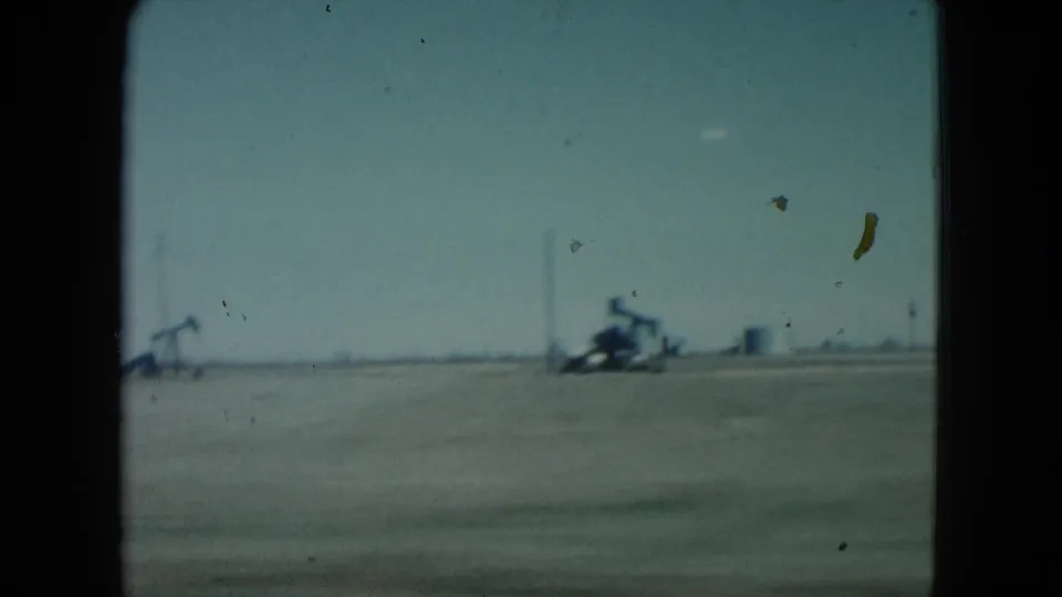 1960 Nevada 8mm frame showing flat desert terrain, visible pumpjacks and distant structures along the horizon, with film dust across the sky.