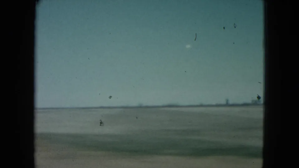 1960 Nevada 8mm frame with a broad flat landscape, pale blue sky, and a dark vehicle-window edge in the foreground.