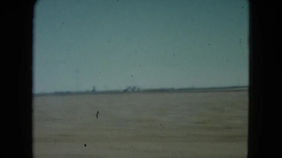 1960 Nevada desert view from a moving vehicle, showing flat open ground, pale sky, and faint structures on the horizon.