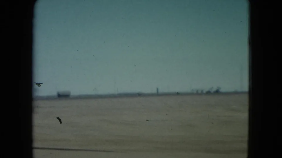 Sparse Nevada desert landscape in 1960, with low horizon structures and a clear sky seen from a passing vehicle.