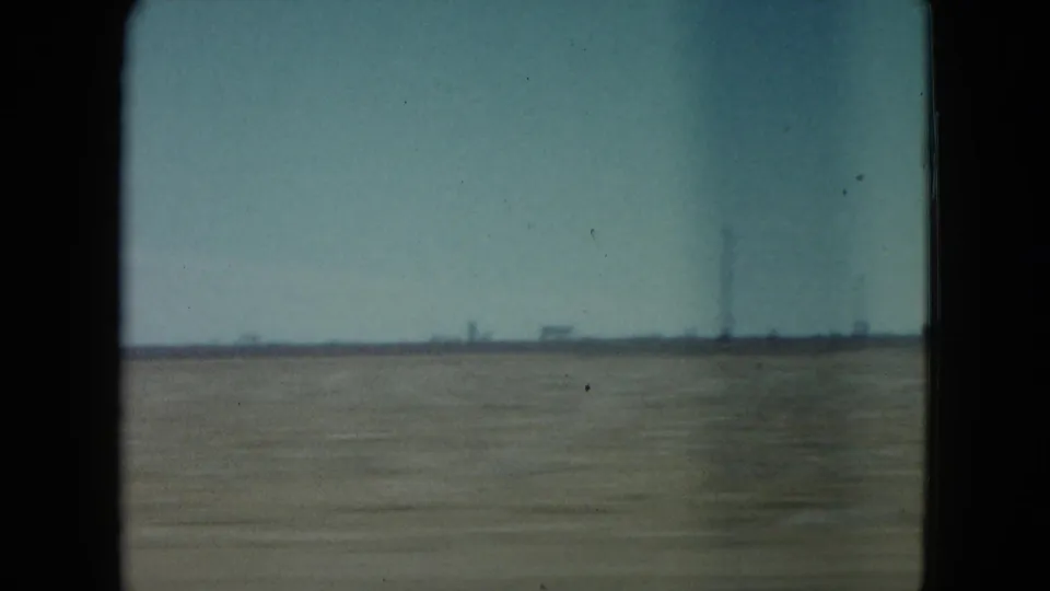 Nevada desert horizon seen from a moving car, with a tall distant pole or stack and low structures under a pale sky.