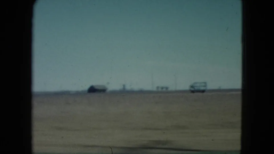 Moving 8mm view across a flat Nevada field, with a distant car and low horizon objects softened by motion and focus.