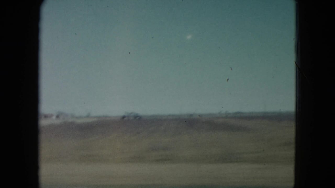 1960 Nevada 8mm frame from a moving vehicle showing a flat field, pale sky, and distant low structures.