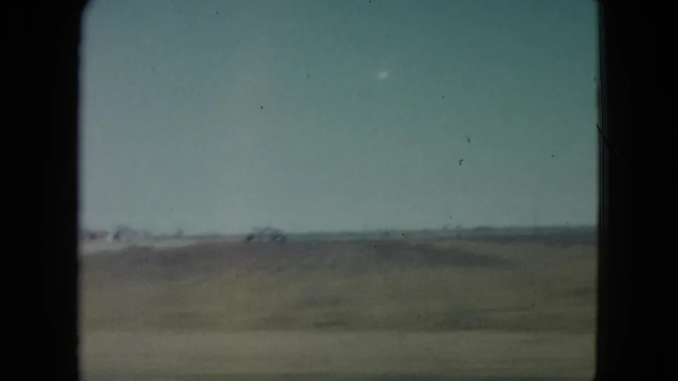 1960 Nevada 8mm frame from a moving vehicle showing a flat field, pale sky, and distant low structures.