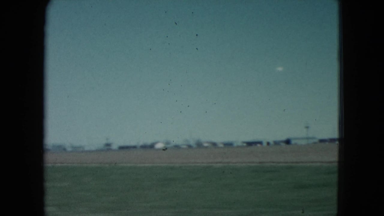 1960 Nevada roadside frame showing green field, distant low structures, and a telephone pole under a pale sky.