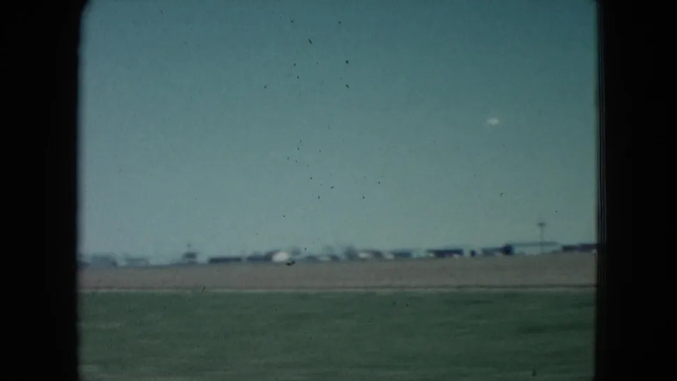 1960 Nevada roadside frame showing green field, distant low structures, and a telephone pole under a pale sky.