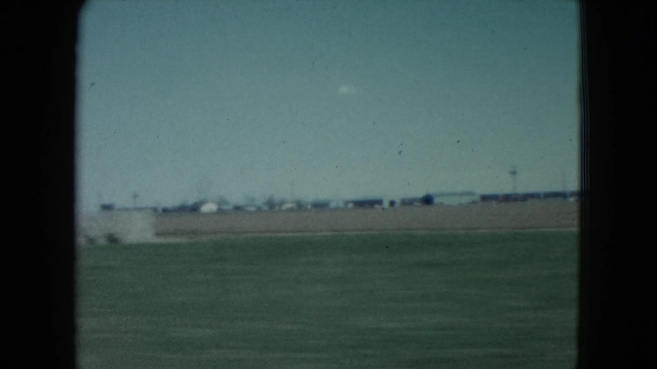 1960 Nevada train-window view across flat arid ground, distant structures, and a pale blue sky, framed by dark film and window edges.