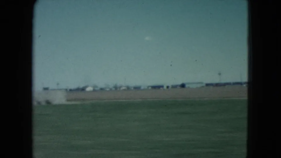 1960 Nevada train-window view across flat arid ground, distant structures, and a pale blue sky, framed by dark film and window edges.