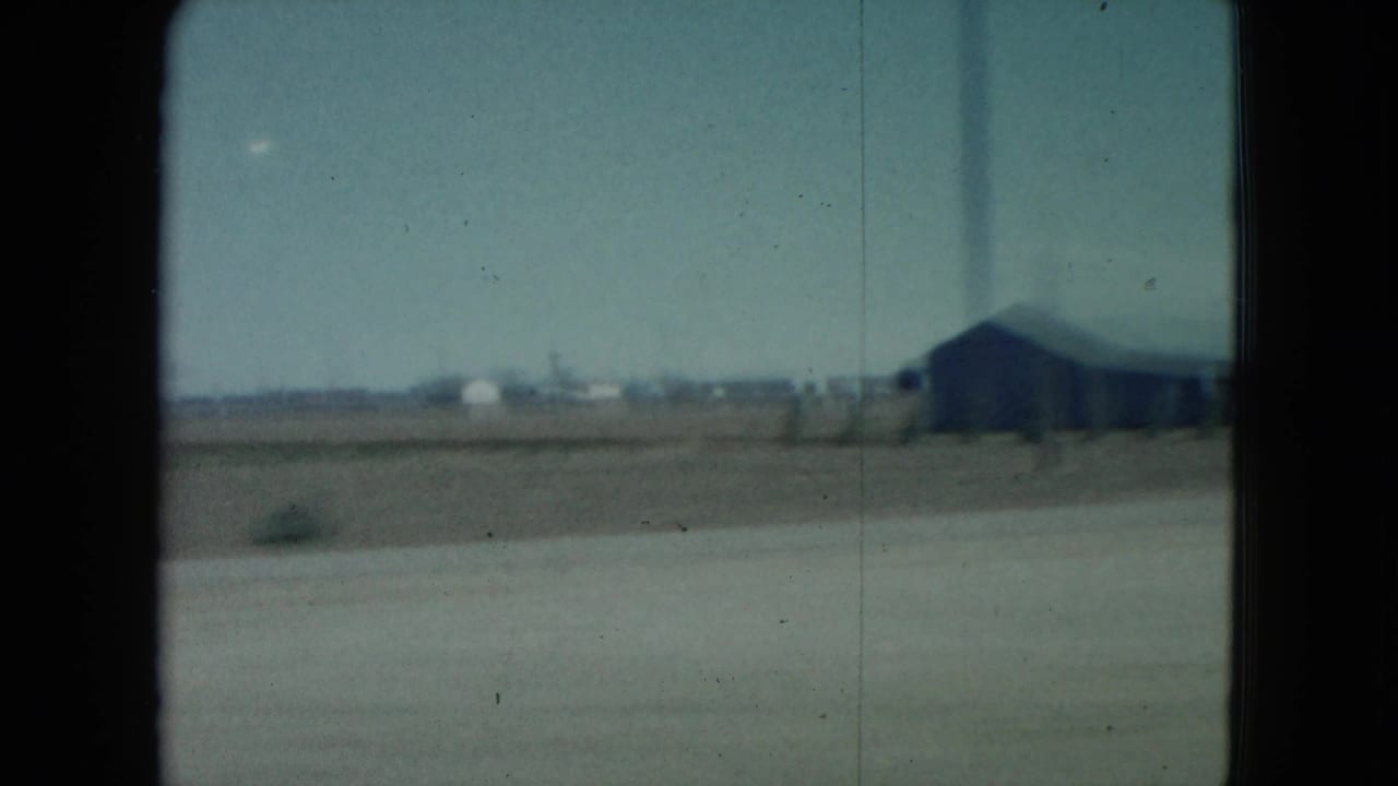 Trackside landscape with a large dark shed-like structure in the distance and window framing at the sides.