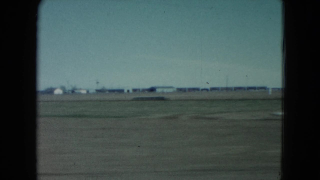 1960 Nevada view from a moving train with open ground in the foreground, distant low buildings, utility poles, and a washed blue sky.