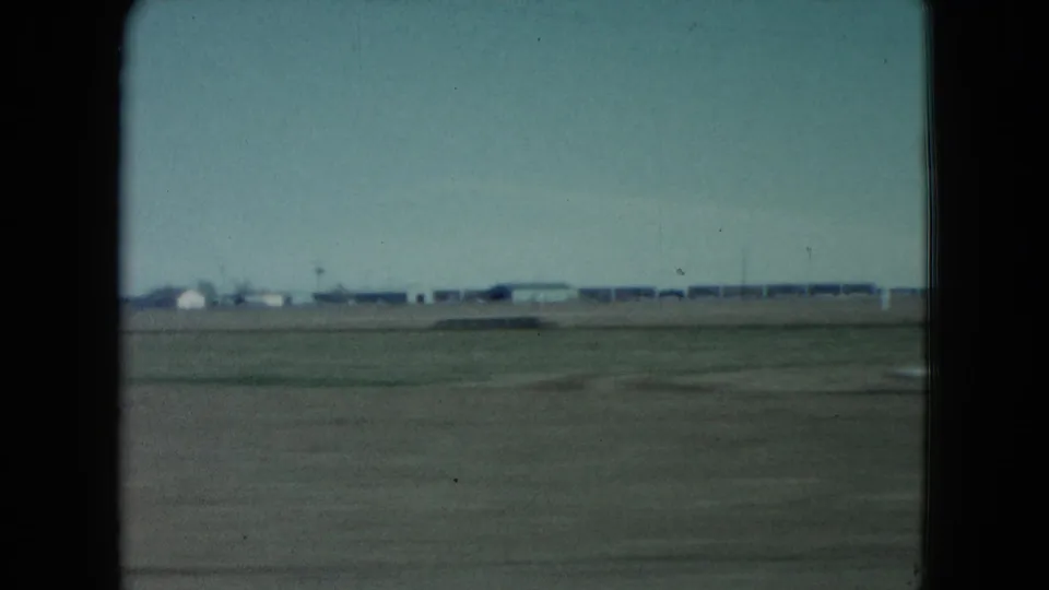 1960 Nevada view from a moving train with open ground in the foreground, distant low buildings, utility poles, and a washed blue sky.