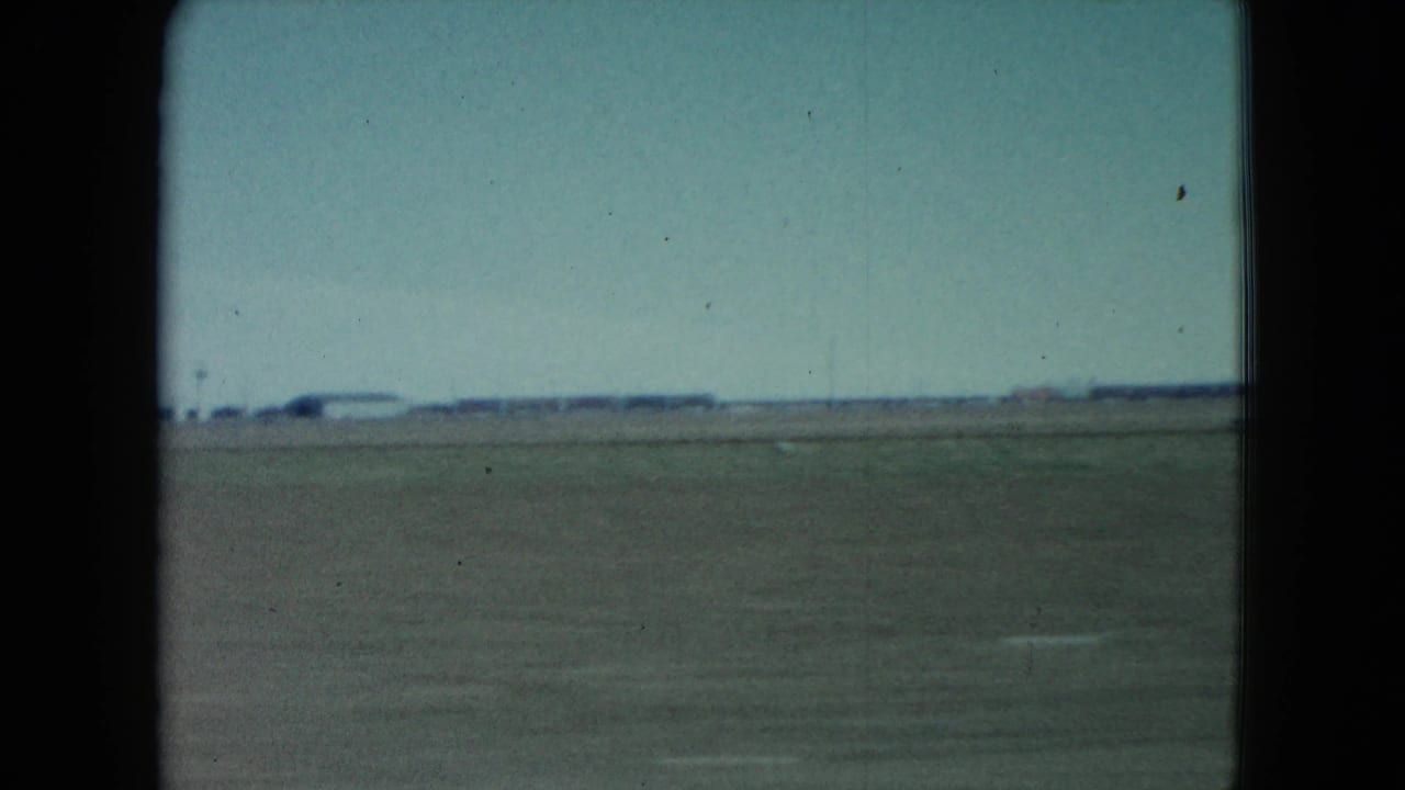 1960 Nevada train-window view over broad flat terrain toward distant low structures, with dark frame edges and visible 8mm grain.