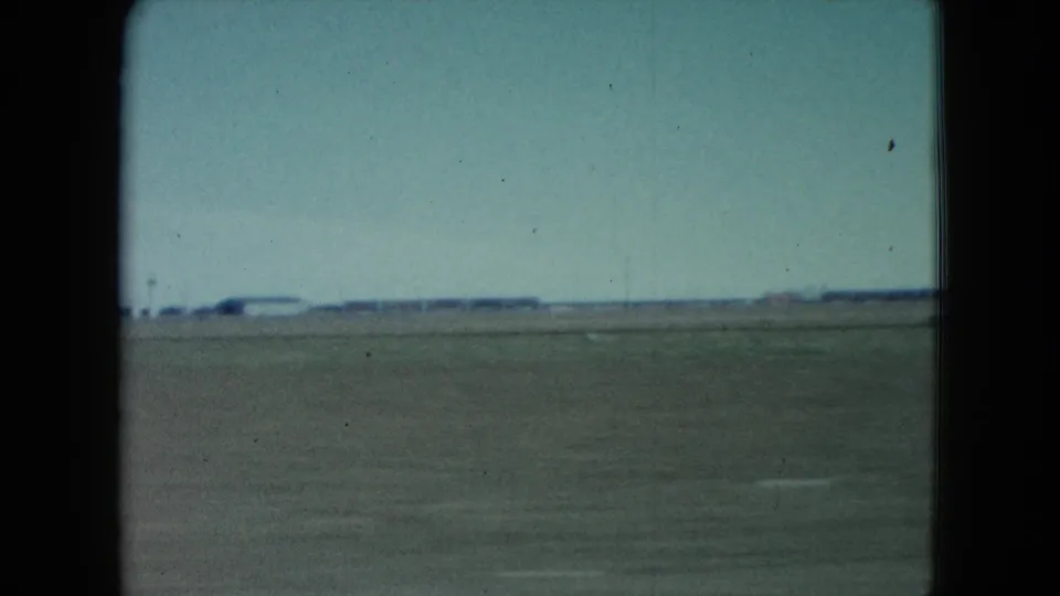 1960 Nevada train-window view over broad flat terrain toward distant low structures, with dark frame edges and visible 8mm grain.