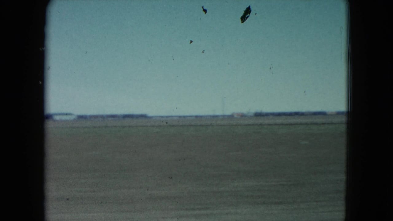 1960 rural Nevada farmland appears beyond a moving car window, with open fields, low horizon, pale sky, and visible 8mm frame edges.