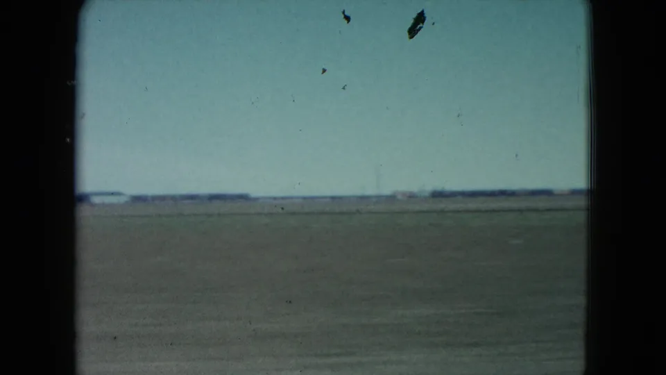 1960 rural Nevada farmland appears beyond a moving car window, with open fields, low horizon, pale sky, and visible 8mm frame edges.