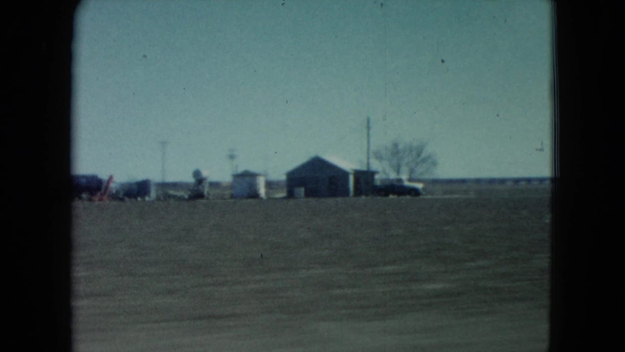 Small farm buildings, utility poles, and a parked vehicle appear in the distance across open rural Nevada fields in this 1960 8mm frame.