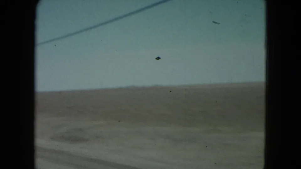 1960 Nevada road-travel frame showing flat arid land, pale sky, and car-window framing from a moving vehicle.