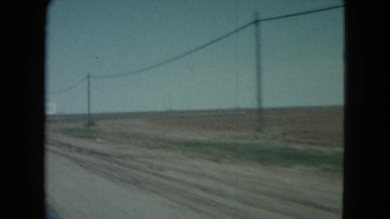 1960 Nevada home-movie frame showing a dirt road, utility poles, wires, and broad open land from a car window.