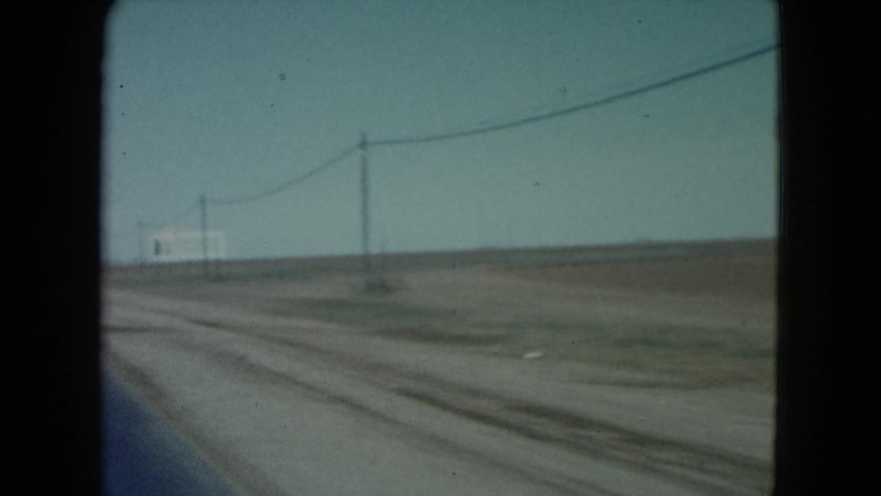 Forward view from a moving vehicle beside a flat desert road with utility poles and overhead wires.