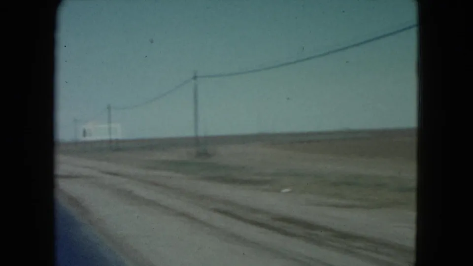 1960 Nevada 8mm frame showing a forward-moving road view across flat desert terrain with utility poles, wires, and a distant roadside sign.
