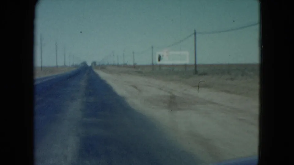 1960 Nevada road footage shows a straight two-lane highway stretching into the distance, with dirt shoulder, utility poles, wires, and a white sign.