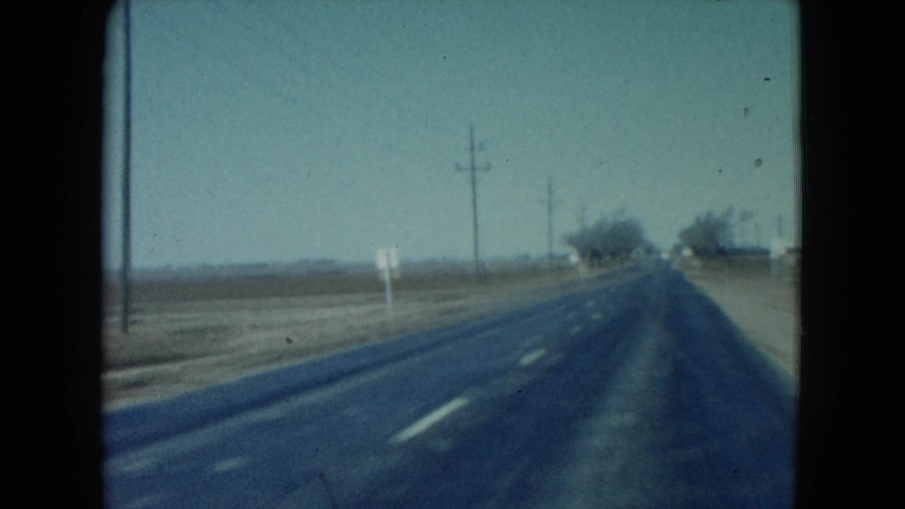 1960 Nevada highway frame shows lane markings, a dirt shoulder, utility poles, small roadside signs, and distant trees along the road.