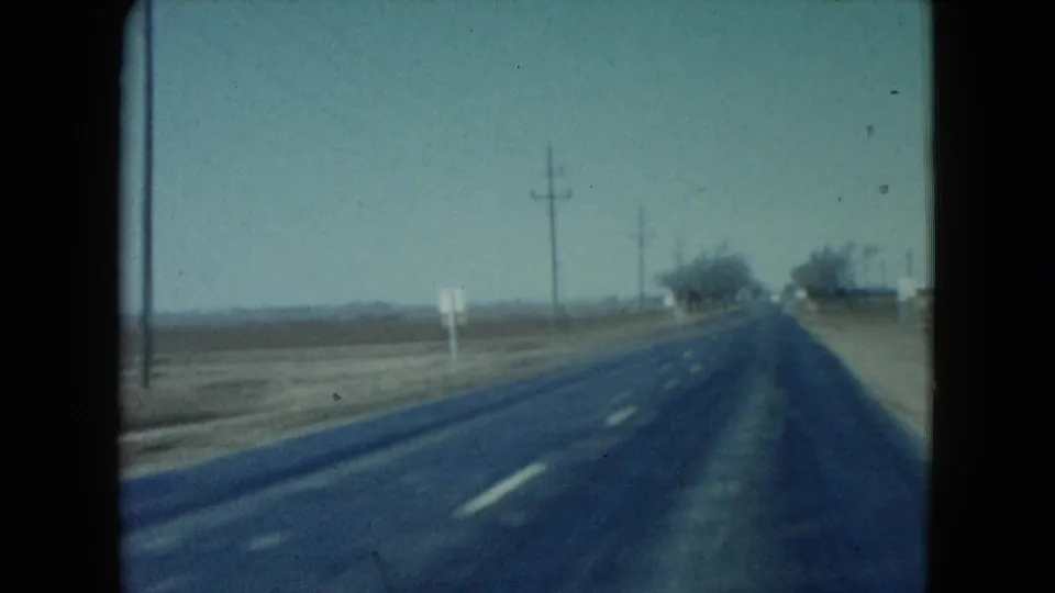 1960 Nevada highway frame shows lane markings, a dirt shoulder, utility poles, small roadside signs, and distant trees along the road.