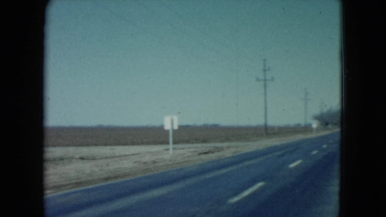 1960 Nevada home-movie frame looking forward along a rural two-lane blacktop road bordered by flat farmland, telephone poles, and a pale sky.