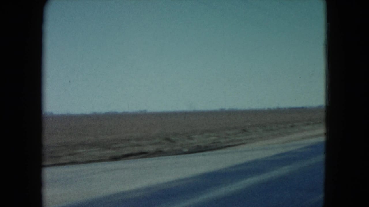 1960 Nevada road-travel frame showing a broad flat field, paved roadside edge, low horizon, and soft motion blur from a moving car.