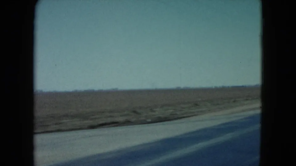 1960 Nevada road-travel frame showing a broad flat field, paved roadside edge, low horizon, and soft motion blur from a moving car.