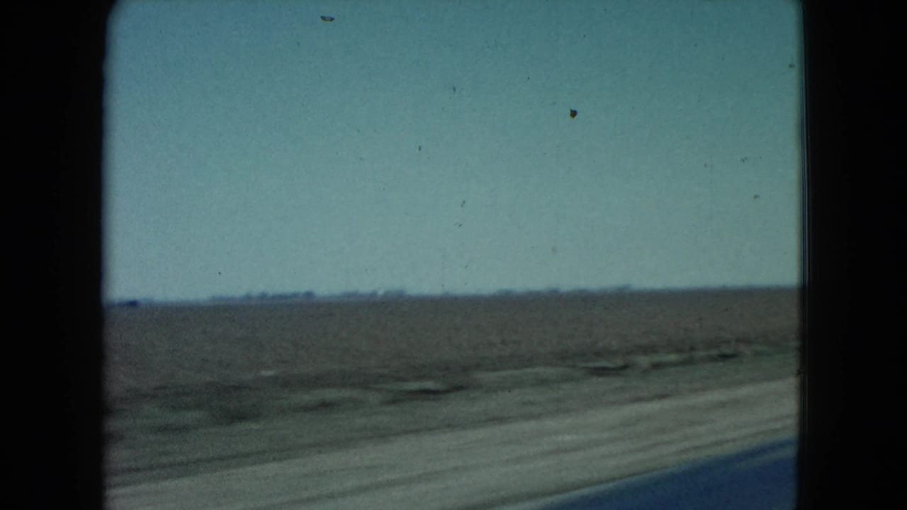 1960 Nevada home-movie frame with open harvested farmland, a low distant horizon, and the blurred edge of the blacktop road in the foreground.
