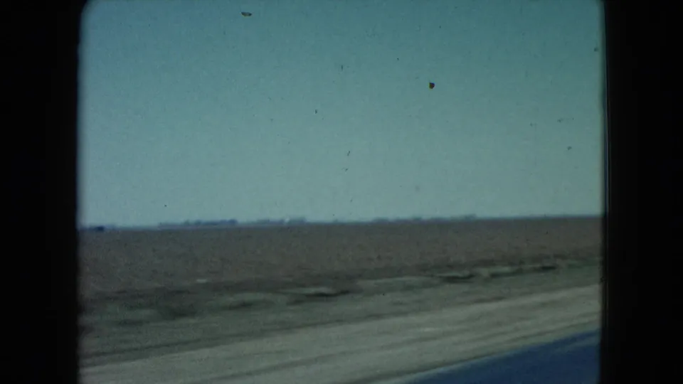 1960 Nevada home-movie frame with open harvested farmland, a low distant horizon, and the blurred edge of the blacktop road in the foreground.