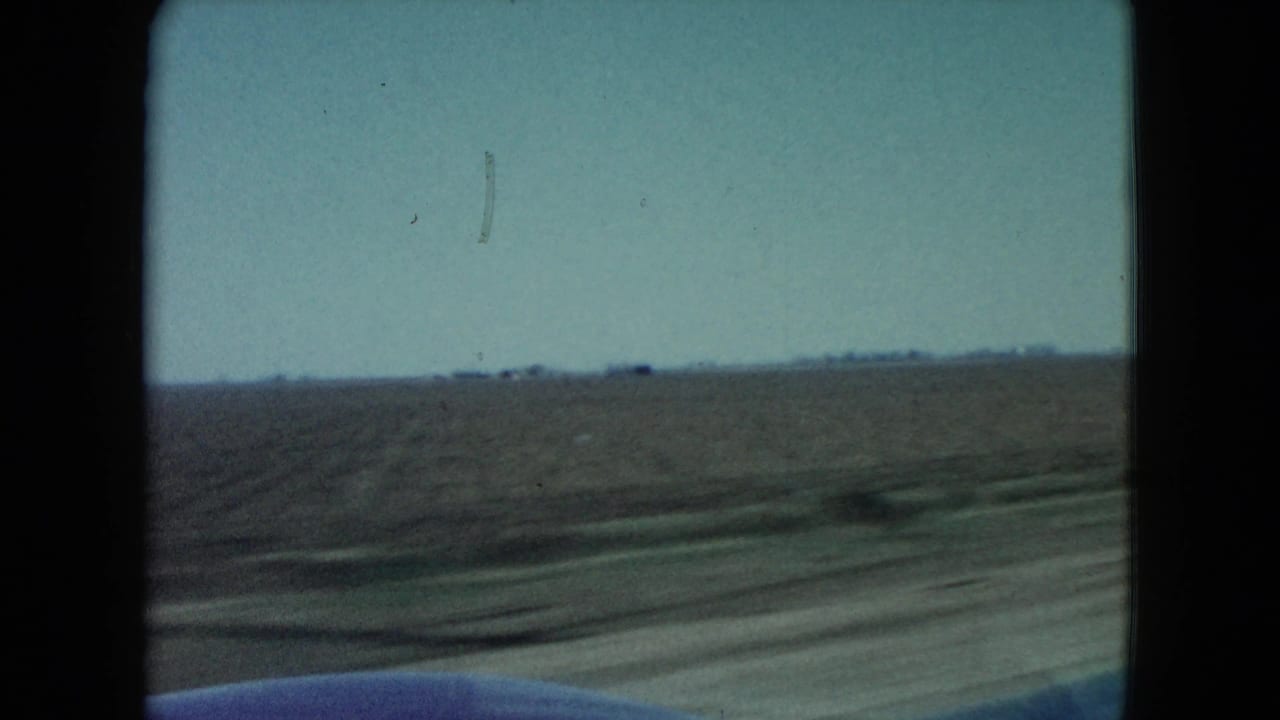 1960 Nevada rural road frame showing flat farmland, a distant low horizon feature, blurred roadside ground, and part of the vehicle at the bottom edge.