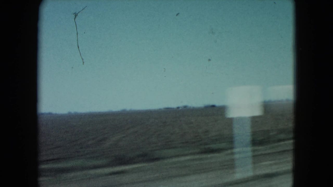 1960 rural Nevada 8mm frame showing flat roadside land, a pale sign, distant horizon, and dark film gate edges during travel.