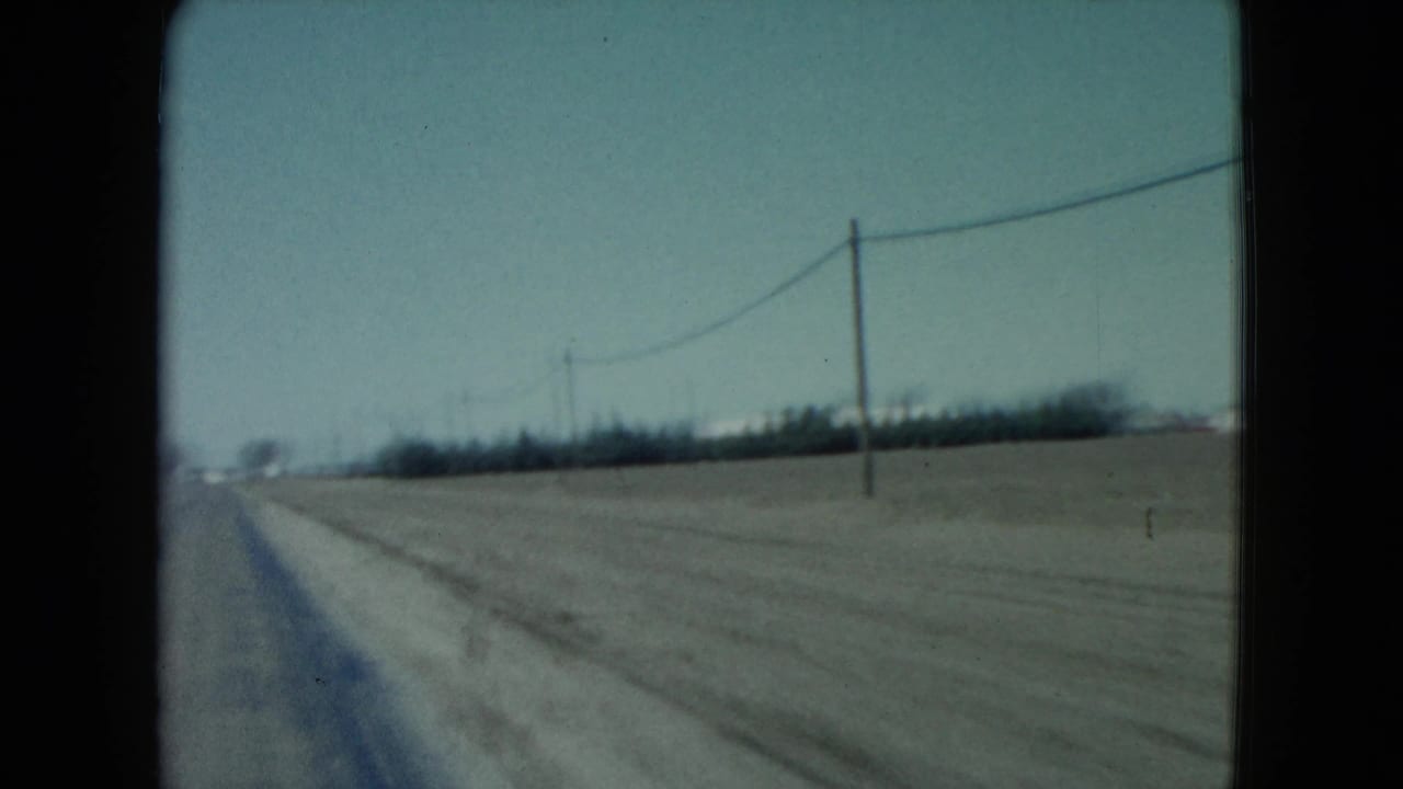 1960 rural Nevada 8mm frame showing the highway edge, open fields, utility poles, wires, and distant trees from a moving vehicle.