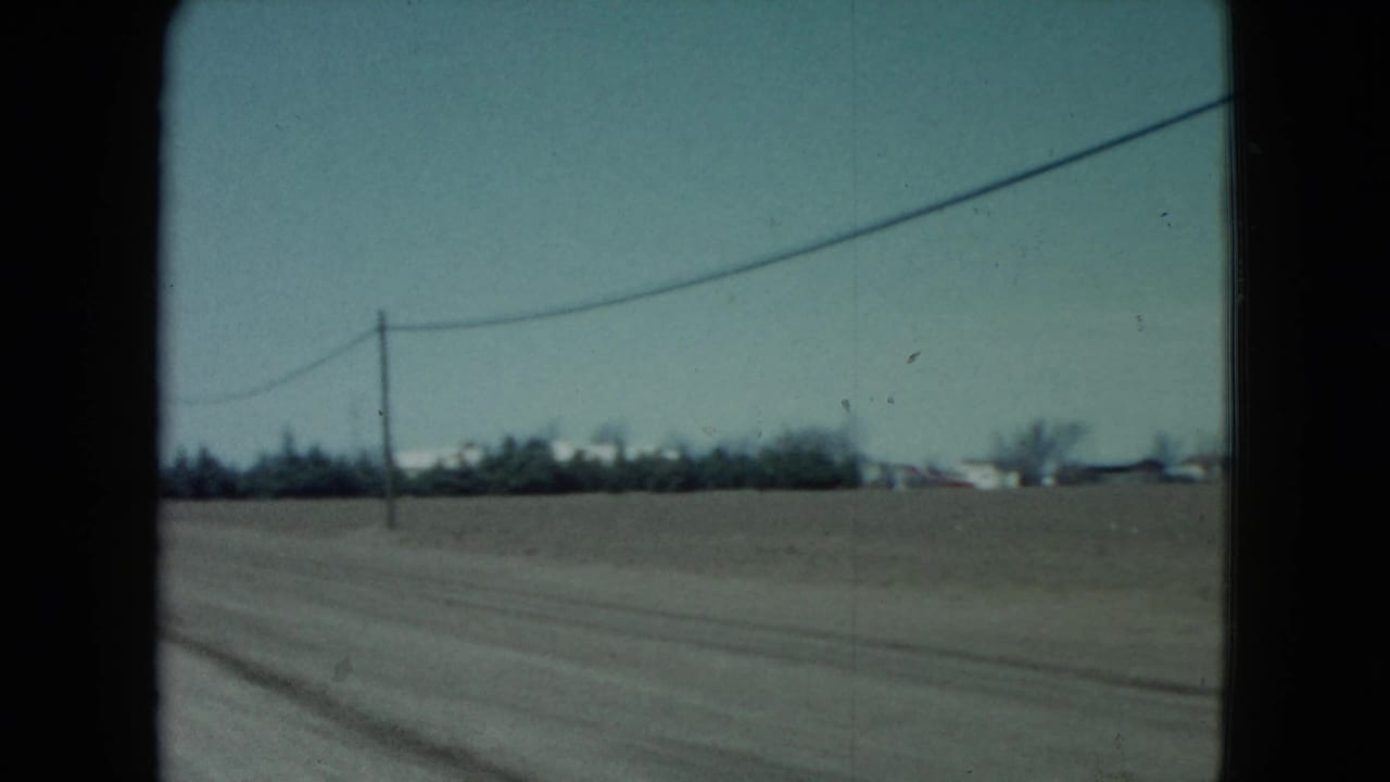 1960 rural Nevada roadside frame with flat fields, a utility line, distant trees, and low buildings glimpsed from a moving vehicle.