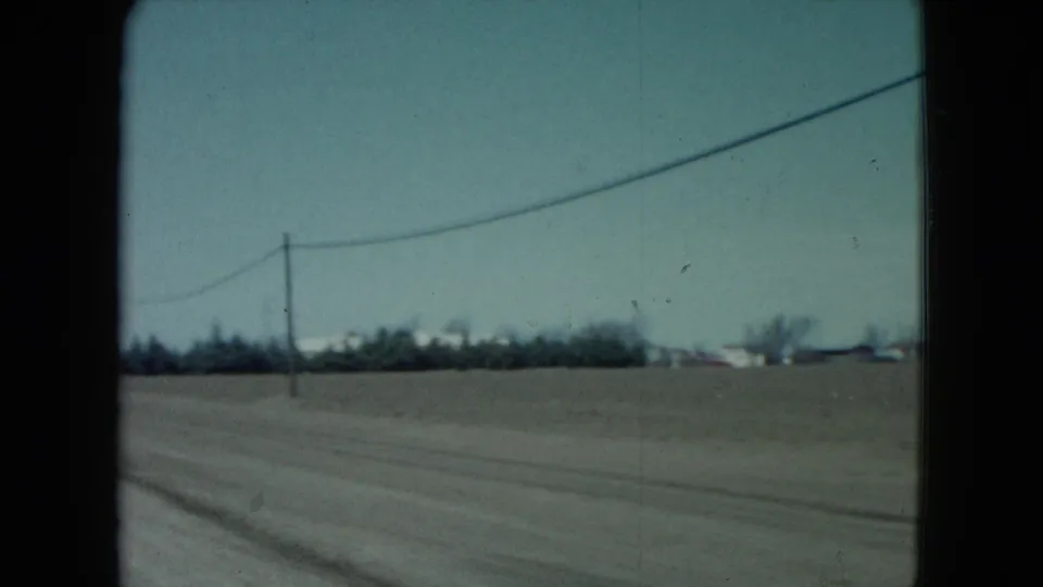 1960 rural Nevada roadside frame with flat fields, a utility line, distant trees, and low buildings glimpsed from a moving vehicle.