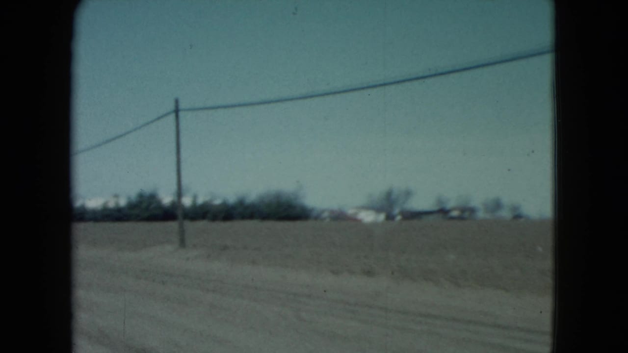 1960 Nevada rural roadside view with flat farmland, a utility pole, overhead line, and small houses on the horizon.